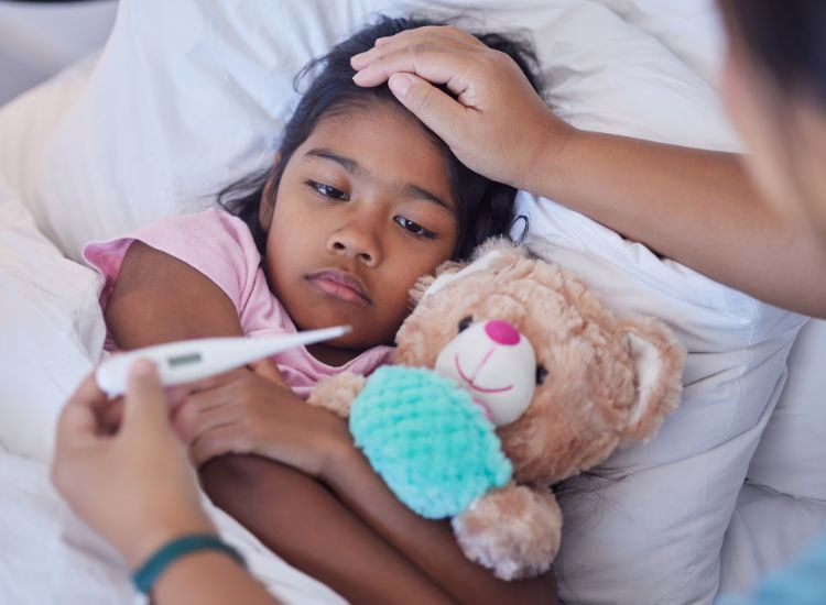 A young girl lies in bed holding a stuffed teddy bear, looking unwell. An adult checks her temperature with a thermometer while gently touching her forehead.