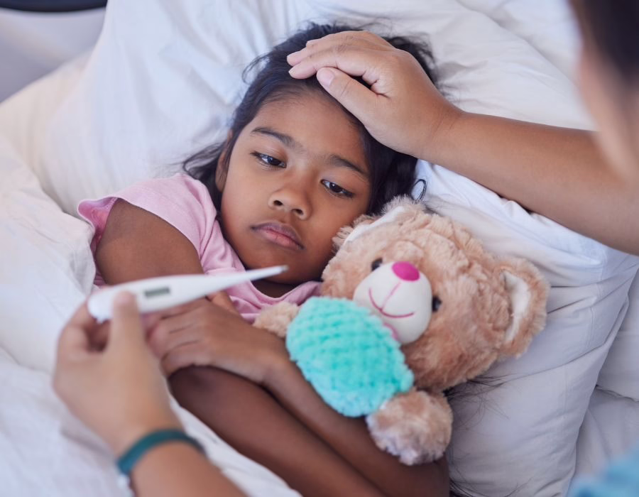 A child holding a teddy bear lying in bed while being checked with a thermometer.